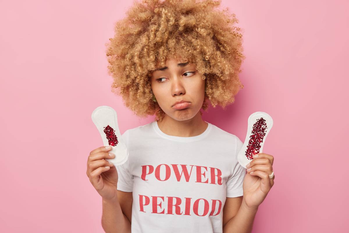Upset curly haired woman has critical days holds sanitary napkins smeared with blood thinks about hygiene protection dressed in casual t shirt isolated over pink background. Gynecological concept