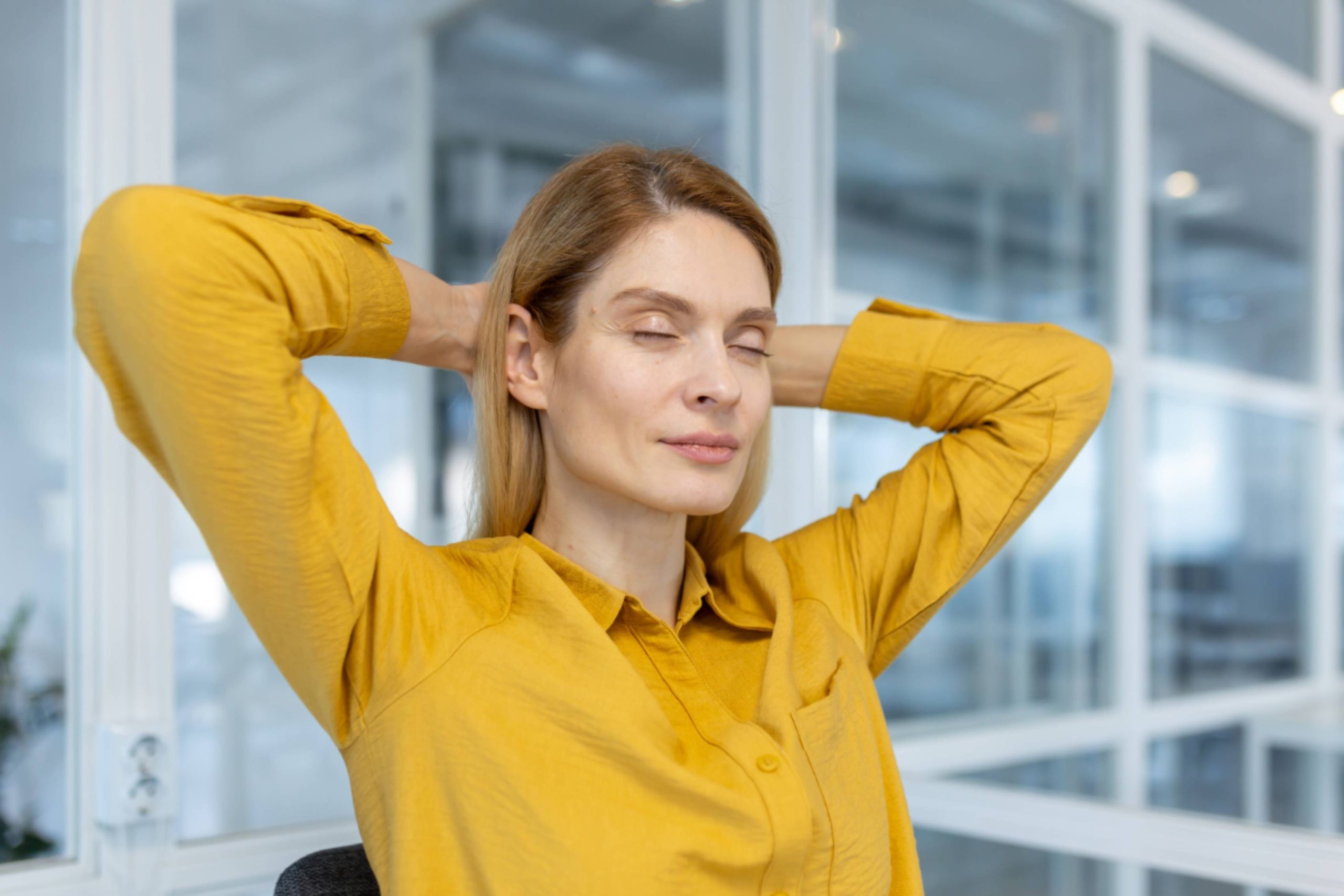 Woman in yellow shirt relaxing with hands behind head in office setting